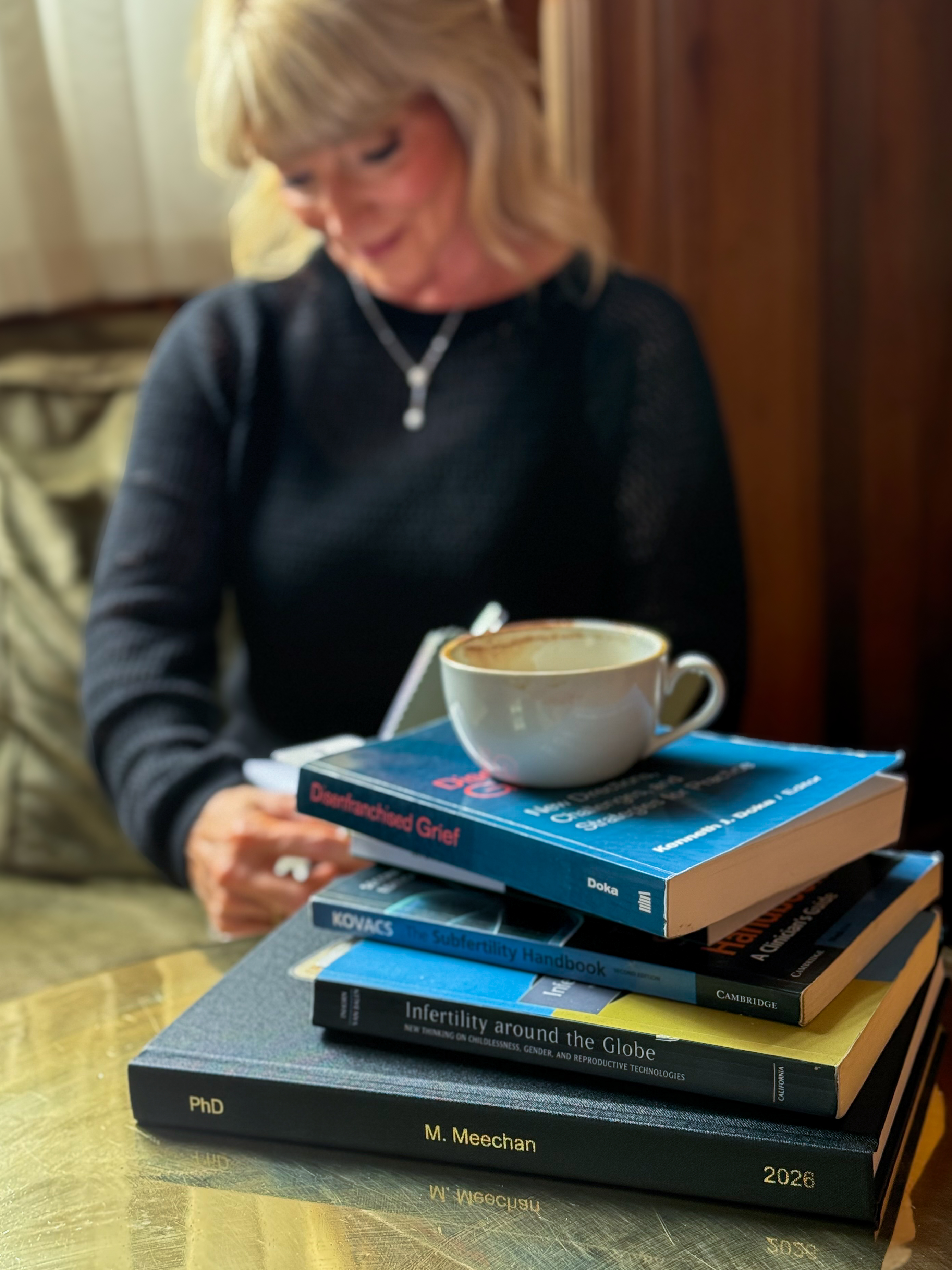 Dr Marie Meechan reading at a table, a cappuccino resting on a stack of her fertility and grief research books.