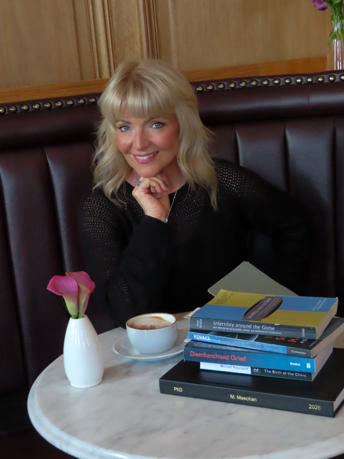 Dr Marie Meechan at a marble cafe table with a cappuccino and a stack of her fertility and grief research books.