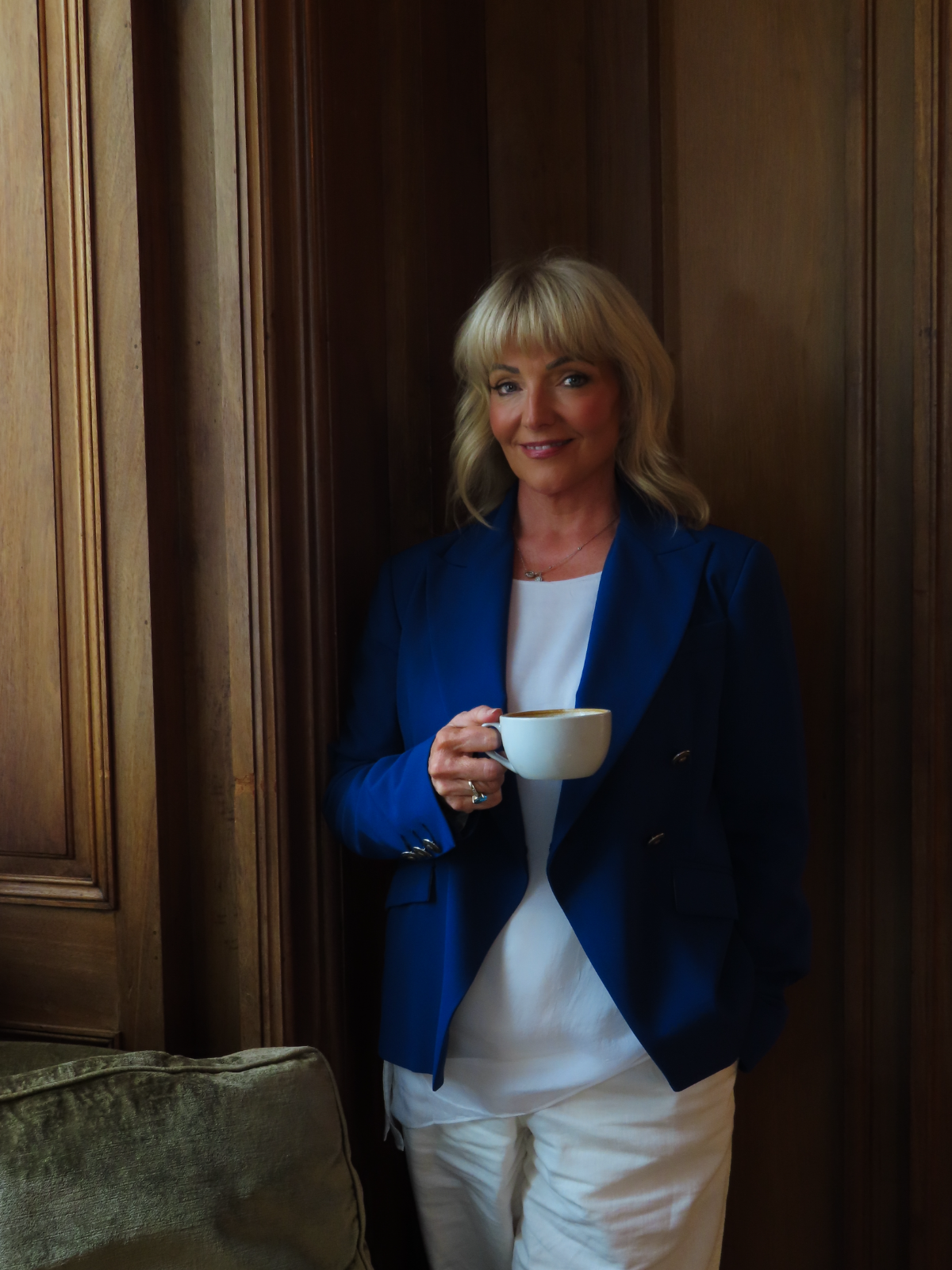 Dr Marie Meechan standing in a softly lit, wood-panelled room, holding a cup of coffee and smiling gently.