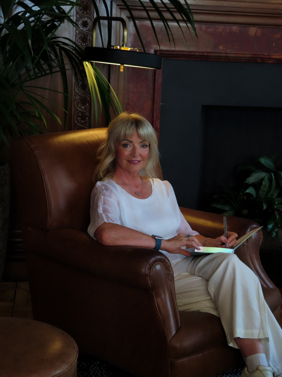 Dr Marie Meechan sitting in a warm leather armchair, writing in a notebook beside a softly glowing lamp and palm.