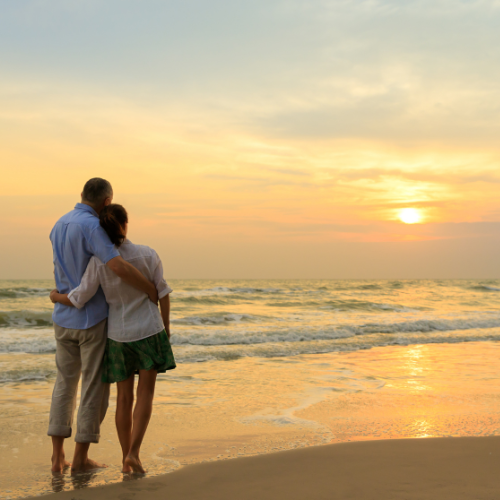 A couple standing together on a beach at sunset, looking out toward the horizon.