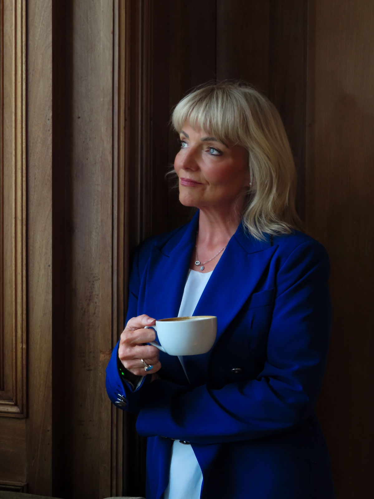 Dr Marie Meechan in a cobalt blazer, holding a coffee and looking out of a window in warm wood-panelled light.
