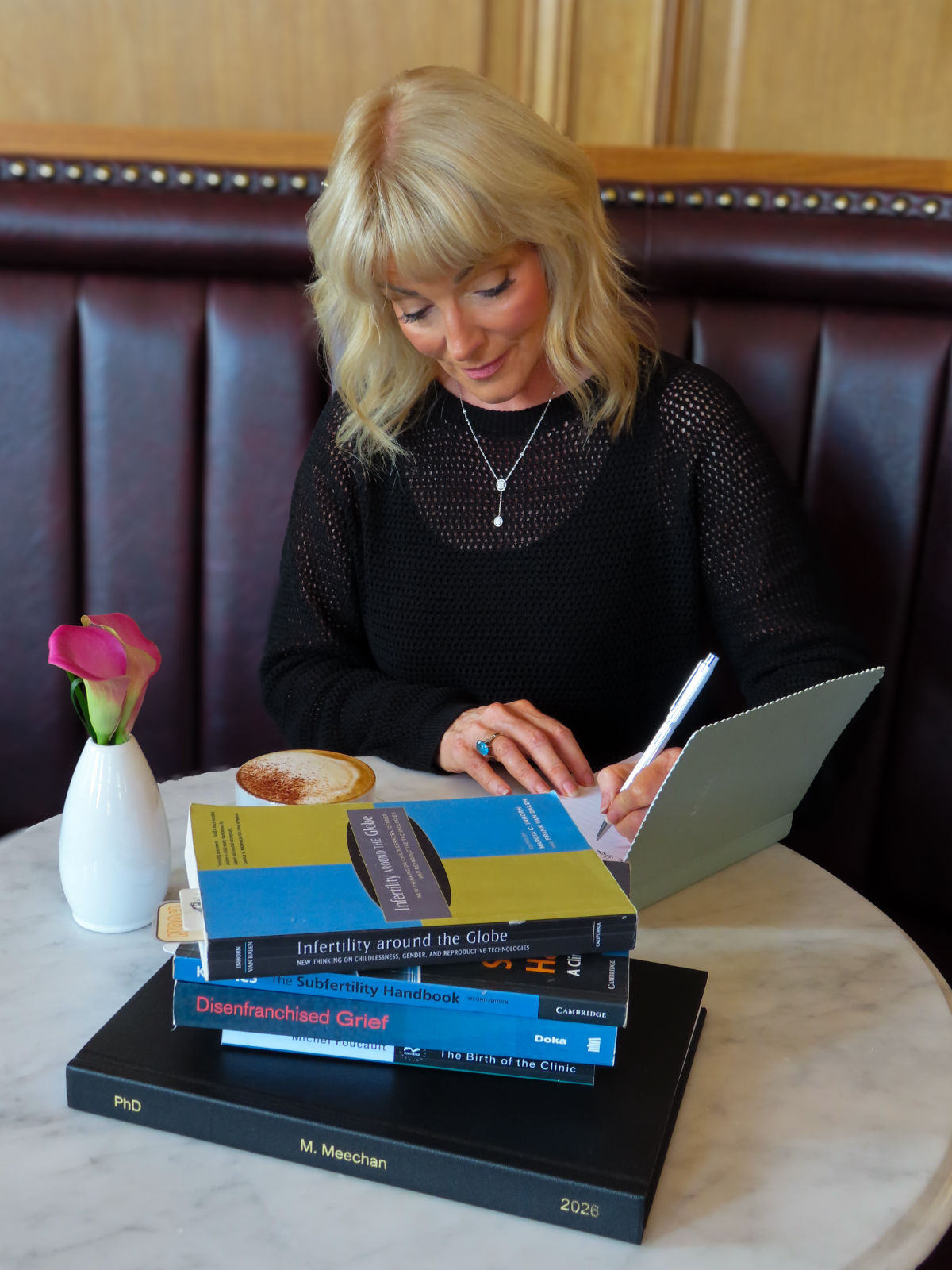 Dr Marie Meechan writing in a notebook at a marble cafe table, a cappuccino and her fertility and grief research books beside her.